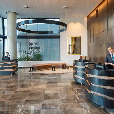 מלון Pullman Brisbane Airport מראה חיצוני תמונה The image shows a modern hotel lobby. There are two reception desks made of dark-colored materials, and staff members are attending to guests at these desks. The lobby features large windows that allow natural light to fill the space, and there are p